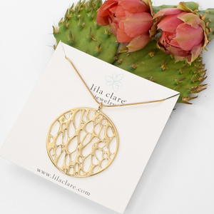 Image of a gold large framed circle cactus skeleton necklace on white card leaning on prickly pear cactus pad and flowers.