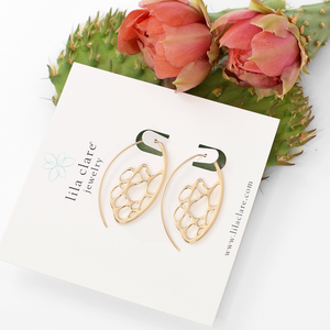 Image of a gold oval cactus skeleton hoop earrings on white card leaning on prickly pear cactus pad and flowers.