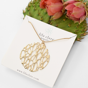 Image of a gold large circle cactus skeleton necklace on white card leaning on prickly pear cactus pad and flowers.